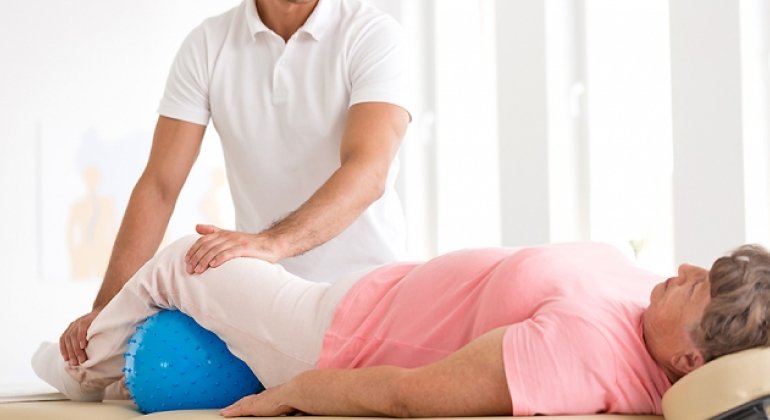A physiotherapist assisting an elderly woman with a leg exercise during a home physiotherapy session. The woman is lying on her back with her legs bent over a large blue therapy ball. The therapist is supporting and guiding her leg movements to ensure proper technique and maximize benefits. The setting is a bright and comfortable home environment.
