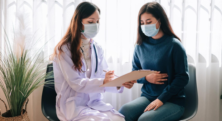 Female doctor wearing a mask consulting a patient who is holding her stomach in discomfort, possibly discussing digestive issues or symptoms of navel displacement.