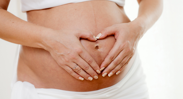 Close-up of a pregnant woman gently forming a heart shape with her hands over her belly button, symbolizing love, care, and connection during pregnancy.