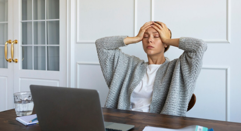 Woman sitting at a desk with a laptop, holding her head with both hands, appearing stressed or fatigued, possibly experiencing headache, dizziness, or mental exhaustion.