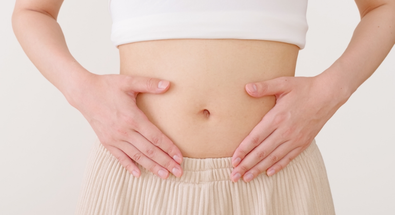 A person wearing a white top and beige pants places their hands on either side of their lower abdomen, focusing on the navel area, symbolizing abdominal or digestive health.