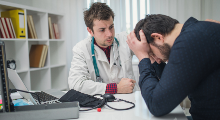 A doctor attentively listens to a distressed male patient who is holding his head in frustration, representing the emotional and physical impact of undiagnosed health issues such as navel displacement.