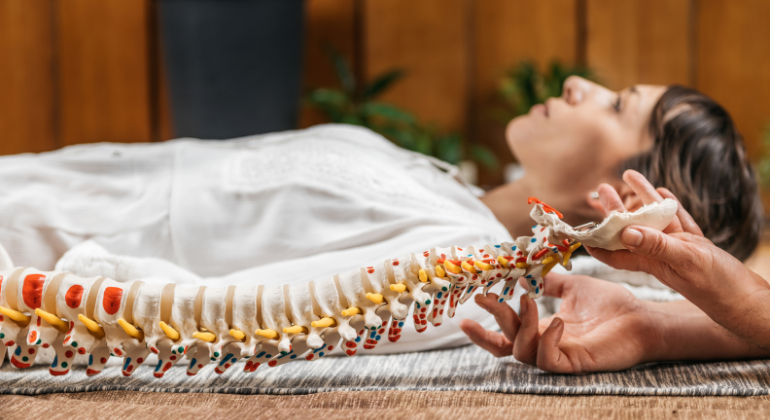 A person lying on their back during a therapy session while a practitioner holds a detailed spine model, illustrating how chiropractic care may support natural correction of navel displacement and spinal alignment.