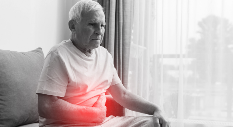  An elderly man sitting on a couch holds his abdomen with a pained expression, with a red highlighted area over his stomach, indicating abdominal pain or digestive discomfort.