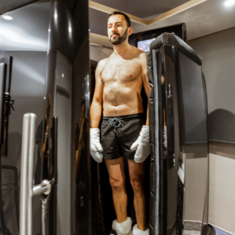 A man is standing inside a cryotherapy chamber. He is shirtless, wearing black shorts, white gloves, and socks, with his eyes closed, appearing calm and relaxed as the treatment begins. The chamber is sleek and modern, with a digital screen on the side displaying settings and controls. The room has a minimalist design with neutral tones, enhancing the clinical yet comfortable atmosphere.